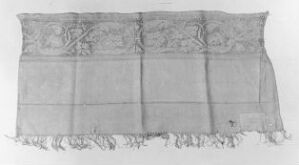  Monochrome photograph of a rectangular fabric with an ornate border and fringe details, displayed against a light background.