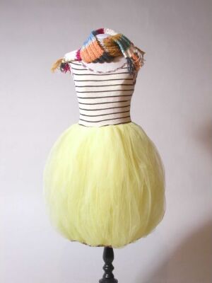  A mannequin draped in a striped sleeveless top in white and faded red and a pastel yellow tutu skirt, topped with a colorful, mixed-hue headpiece against a light background. Artist name and title unknown.