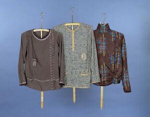  Three garments displayed against a blue background; from left, a deep brown top with white stitching, a grey marled cardigan with a yellow zippered front, and a dark brown shirt with a colorful plaid pattern.