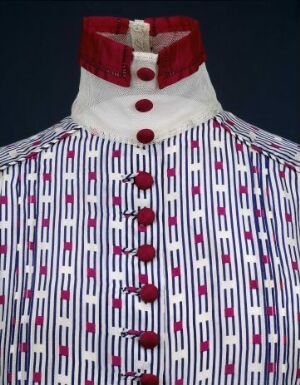  A close-up of a shirt or jacket with a high, velvet maroon collar and a white chest area adorned with vertical navy blue and burgundy stripes, interspersed with small purple Eiffel Tower motifs, and closed with round crimson buttons.