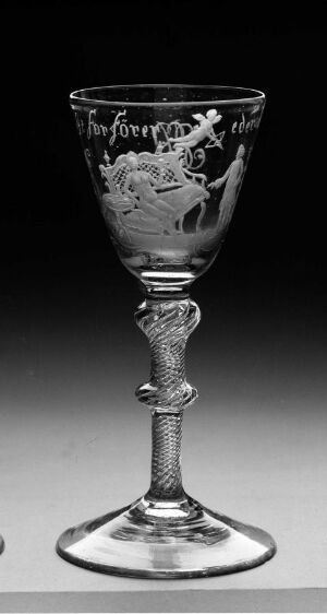  A grayscale image of a clear glass goblet named "Nøgne Jomfruer," featuring an etched design on the bowl and a decorative stem that mimics a rope or beaded pattern, set against a neutral gray background.