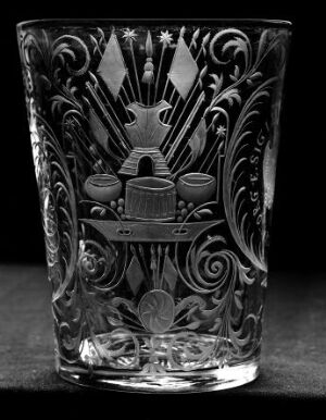  A black-and-white photograph of a clear glass with intricate etched patterns, featuring a central emblem with a sunburst top, wing-like shapes, two barrels, and surrounding leafy swirls, against a black background.