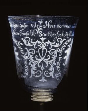  An engraved glass by Nøstetangen glassverk with a flaring bowl and a short stem on a circular base. The clear glass features white, detailed baroque engravings, including text and a central emblem, starkly contrasting against a dark background.