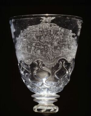  An intricately etched or engraved glass goblet with a detailed floral pattern and symmetrical bird-like designs in the center against a dark background, showcasing the transparent and reflective qualities of the glass.