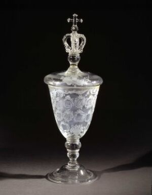  An intricately designed glass or crystal chalice with a detailed etched pattern on its bowl, adorned with a lid topped by a cross sitting on a crown, all against a dark background.