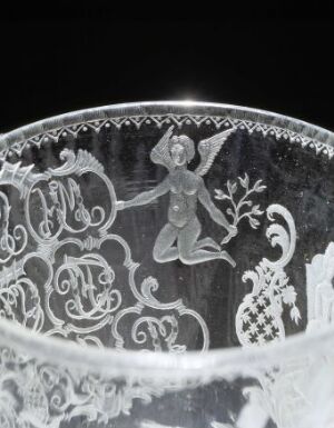  Close-up of a clear glass item titled "Postmester Pind-pokalen" by Hurdal glassverk with frosted engraved decor showcasing a cherubic figure amidst intricate floral patterns and scrollwork on a black background.