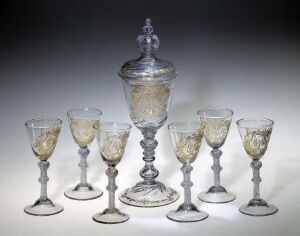  A set of exquisite glass tableware titled "Arctander-pokalen," designed by Johan A. Becker, comprising a large centerpiece goblet with engraved and gilded decorations and a lid with a finial, alongside six smaller matching wine glasses, all displayed against a neutral background. The set embodies a blend of clarity and ornamental finesse, with golden motifs enhancing the elegance of the blown glass pieces.