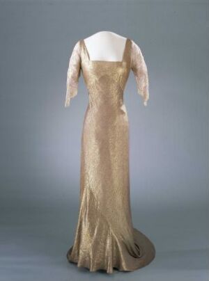  A vintage champagne beige evening gown with off-shoulder neckline and sheer long sleeves featuring subtle embroidery, displayed on a mannequin against a gray background. The dress fits snugly at the bodice and hips before flaring out to a gently trailing skirt, capturing the elegance of early 20th-century fashion.