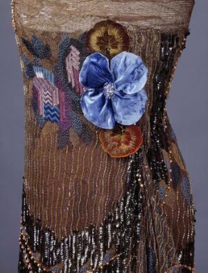  A close-up of a textured fabric featuring a large blue satin flower, a golden to coppery circular embellishment, and a brown and metallic striped background with additional patterned fabric details, suggesting a piece of textile art or a detailed section of a garment.