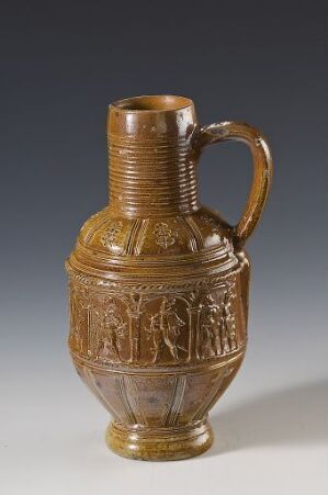  A historical honey-brown ceramic jug with a ribbed neck and handle, featuring intricate relief scenes on its body, displayed against a neutral gray background.