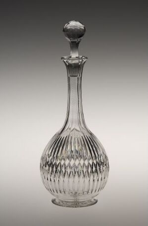  An elegant, transparent glass decanter with a faceted stopper and a vertically ridged body, displayed against a graduated gray background.