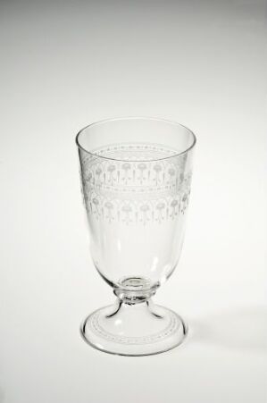  A clear, etched glass goblet with a decorative lace-like pattern around the upper half, short stem with a single knop, and a stable base, set against a light grey background. Artist name and title are unknown.