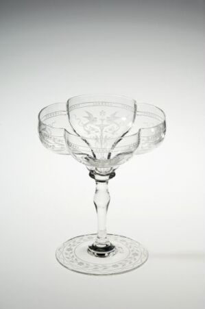 
 An elegant clear glass with an etched floral or vine-like design on the bowl and the base, which has a slender stem with a decorative element, set against a soft white to light grey gradient background.