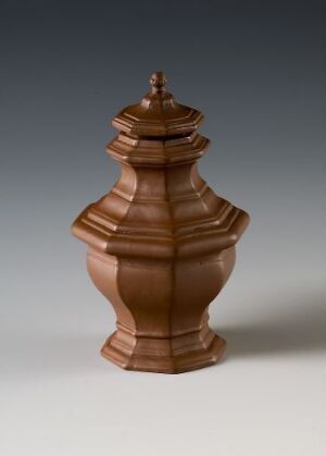  A polished brown decorative urn with concentric ridges, standing on a small base and topped with a pointed finial, set against a soft gray background.