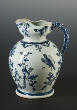  A white ceramic pitcher with intricate blue floral and faunal motifs on a dark grey background, featuring a fluted rim, a swooping spout, and a curved handle.