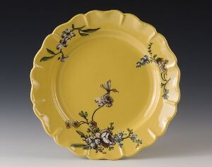  An elegant ceramic plate with scalloped edges, painted with delicate floral patterns on a buttery yellow background. The artist's name and the title of the piece are unknown.