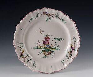  An antique-looking decorative plate with scalloped edges featuring a soft pastel-colored floral border with an Oriental scene at the center, portraying two figures amidst greenery and flowers.