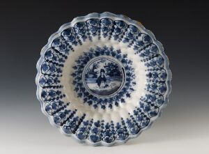  An antique plate with intricate blue and white patterns, featuring a central pastoral scene and scalloped edges against a plain grey background.