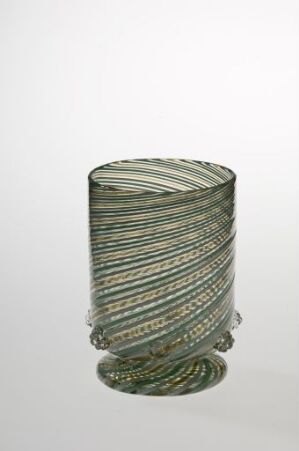  A cylindrical glass object with a swirling pattern of green, brown, and off-white stripes against a light backdrop. Artist name and title are unknown.