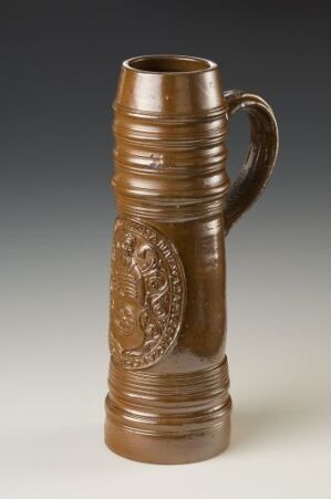  A tall, cylindrical brown stoneware beer stein with horizontal ridges and a raised coat of arms on the front, created by Hubert Schiffer and titled "Mainz' byvåpen." The stein is set against a background that fades from light to dark.