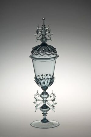  An intricate clear glass goblet with a blueish tint, featuring a metal crown-like cap and decorative metal arms, supported by a simple stem on a flat base, set against a gradient gray background. Artist name and title are unknown.