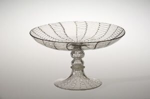  A clear glass compote with intricate patterns, standing on a translucent pedestal with a decorative base, against a softly shaded background.