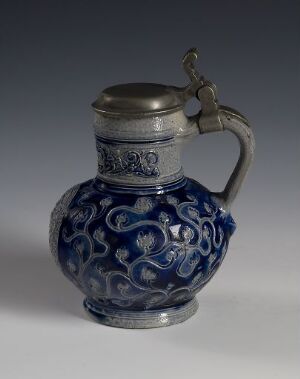  An ornate jug with a cobalt blue ceramic body featuring white floral patterns and a metallic (pewter or silver) neck and handle, set against a graduated grey background.