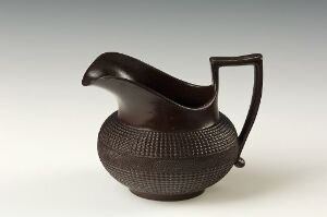  A dark brown ceramic pitcher with a spherical, textured body, elegant curved spout and a flat, rectangular handle on a light neutral background. The artist's name and title of the work are unknown.