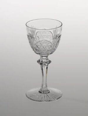  A clear crystal wine glass titled "Hjørdis" by Hadeland Glassverk is displayed against a soft gray background, showcasing intricate cut designs on its bowl and base, reflecting the careful artisanship of design and tableware.