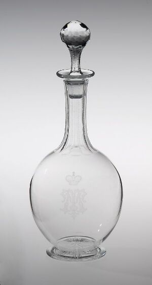  A clear glass decanter with an engraved crest, attributed to Hadeland Glassverk, featuring a faceted stopper and a rounded body on a graduated white to gray background.