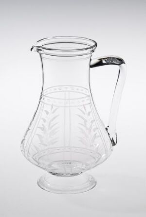  A clear glass pitcher with an etched leaf pattern on the lower half and a smooth arched handle, showcased against a plain light background. The pitcher reflects gentle studio lighting that accentuates its transparent quality and elegant design.