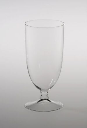  A clear wine glass with a traditional design, featuring a bell-shaped bowl, a straight stem, and a round base, against a light greyish-white background. The glass is empty and the simplicity of the setting underscores the elegance of the glassware.