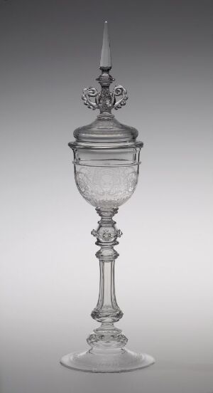  An elegant, translucent glass goblet with an ornate stem and etched designs, topped with a decorative lid and finial, set against a gradient grey background.