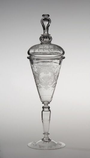  A clear glass goblet or decorative vessel with a conical lid adorned with frosted etchings, featuring an ornamental finial on top, set against a plain light grey background.