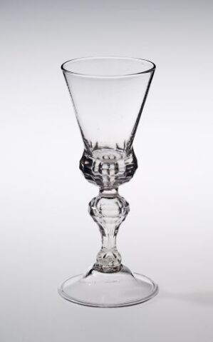  A clear glass wine goblet with an ornate figure in the center of the stem, placed against a light grey background. The goblet features a wide bowl suitable for red wine, a decorative stem with a figure-like element, and a stable, flat foot.