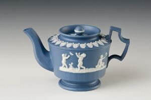  A cobalt blue-glazed teapot with a raised white decorative band featuring dancing figures around the body, a gracefully curved spout, and a swooping handle, against a neutral pale gray background. Artist name and title remain unknown.