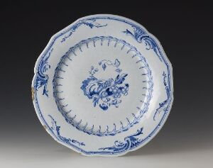  A round faience plate with scalloped edges, featuring a central floral bouquet motif and intricate blue patterns on a white background, titled "Det rehnska mönsteret" by an unknown artist.