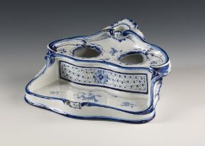  An antique white porcelain inkstand with intricate blue floral and vegetal designs. It has a wavy contour with a cobalt blue rim and two round recesses, possibly for inkwells, on either side of a raised central decorative element.