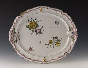  An oval-shaped porcelain platter with scalloped edges highlighted by subtle pink glaze and detailed hand-painted floral designs in yellows, blues, greens, and magenta. The edges feature pale pink ribbon-like decorations.