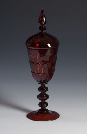  A transparent red faceted glass goblet with a helix stem and a pointed cover on a neutral gray background.