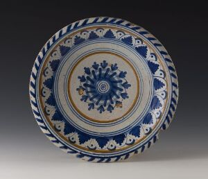  A blue and white ceramic plate with a detailed central design and multiple border patterns, set against a gray background. The design features radial floral motifs and geometric shapes. Artist name and title are unknown.