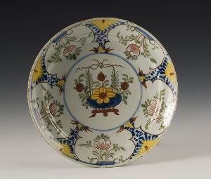  A glazed ceramic plate with a central design of a stylized bouquet in a golden yellow vase, framed by blue scrollwork and bordered with blue, green, yellow, and rusty red floral patterns on a white background.