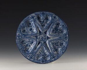  A circular ceramic plate with an ornate eight-pointed star design in cobalt blue on a white background, featuring detailed symmetrical floral patterns and intricate blue glazing. Artist name and title are unknown.