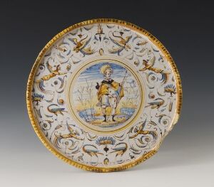  Antique plate with a central illustration of a man in blue and yellow historical attire, surrounded by a decorative rim embellished with cobalt blue and yellow patterns, and a delicate looping design on the outer edge.