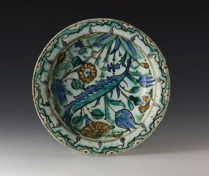  A circular ceramic plate with a detailed botanical design in cobalt blue, turquoise, green, and orange on an off-white background, exhibiting an ornate and colorful aesthetic.