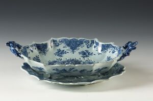  An asymmetrical ceramic bowl with a scalloped edge, painted in a traditional blue and white pattern, resembling historic porcelain designs. The bowl is displayed against a neutral background that contrasts with its vivid blue botanical motifs. The artist's name and title of the work are unknown.