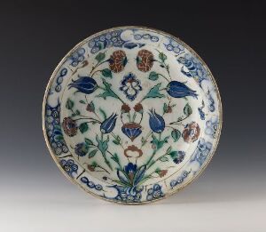  A decorative ceramic plate with a white background and detailed floral pattern in shades of blue, olive green, and rusty brown. The plate has a scalloped edge and sits against a neutral gray background.