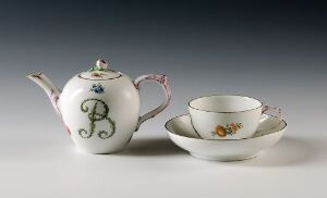  A porcelain tea set comprising a white teapot with a blue monogram and green crown-like detail, a decorated handle, and a flower near the spout is displayed with a matching teacup and saucer featuring floral embellishments on a light grey background.