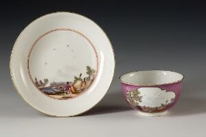  An antique ceramic plate and bowl on a gray background. The plate is white with a golden brown rim and a hand-painted pastoral scene around the edge. The bowl has a magenta exterior and a pastoral scene inside a white oval cartouche, matching the interior of the bowl. Both pieces are delicately painted with muted earthy tones.