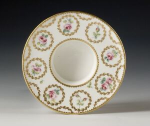  A decorative porcelain saucer with a creamy white background, featuring intricate golden patterns and small floral designs of pink roses with green leaves around the central indentation, all framed by a gold rim, against a dark background. Artist name and title are unknown.